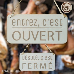 Può includere: Due cartelli in legno con corde appese. Il cartello superiore recita "entrez, c'est OUVERT" e il cartello inferiore recita "désolé, c'est FERMÉ".