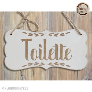 Peut inclure: Une pancarte en bois blanc avec une inscription en brun cursive "Toilette" et un motif de feuilles décoratif. La pancarte a un style rustique et champêtre et est parfaite pour une salle de bain.