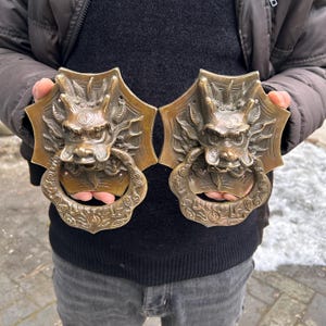 May include: Two antique bronze dragon door knockers. Each knocker features a detailed dragon head with a ring-shaped handle. The bronze has a weathered patina, adding to the vintage aesthetic. The dragon's face is intricately sculpted with sharp teeth and expressive eyes.