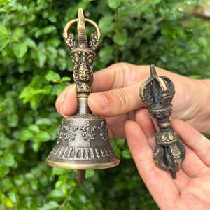 May include: A bronze-coloured metal ritual bell featuring a detailed face at the top of the handle and intricate embossed patterns around the base, accompanied by a smaller, matching bronze-coloured metal vajra scepter with ornate carvings.
