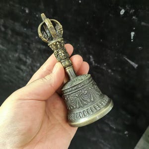 May include: A silver-colored hand bell with intricate carvings and a decorative handle. The bell has a traditional design and is used in religious ceremonies.