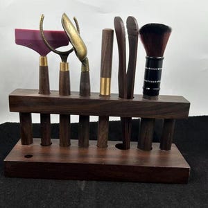 May include: A tea ceremony tool set featuring a wooden stand holding various tools. The tools include a purple spatula, a brass scoop, a wooden tong, and a brush with a black handle. The stand is made of dark brown wood.