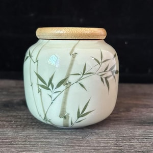 Op de afbeelding: Een kleine, crèmekleurige keramische pot met een bamboe-ontwerp in groen en bruin. De pot heeft een afgeronde vorm en een lichtgekleurde houten deksel. De bamboestengels en bladeren zijn op het oppervlak geschilderd.