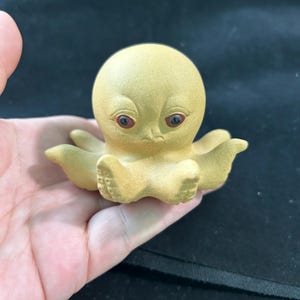 Puede incluir: Pequeña figura de pulpo amarillo con ojos rojos. El pulpo tiene una cabeza redondeada y ocho tentáculos. La figura está hecha de un material liso y tiene un acabado ligeramente brillante. El pulpo está sobre una superficie.
