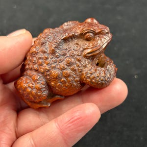 Könnte beinhalten: Eine detaillierte Holzfroschskulptur mit strukturierter Oberfläche und aufwendigen Schnitzereien. Der Frosch hat eine warme braune Farbe und scheint eine Sammlerfigur zu sein. Der Frosch wird in einer Hand gehalten.