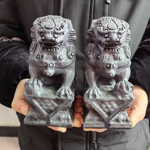 May include: Two gray stone Foo Dog statues. Each statue is in a seated position on a geometric base. The Foo Dogs have detailed features, including a mane, facial features, and paws. The statues are held in front of a dark background.