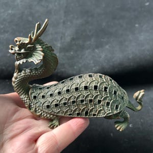 May include: A bronze-toned metal figurine shaped like a mythical dragon-turtle hybrid. The creature features a detailed head with horns and a curved neck, while its shell is composed of an intricate, openwork pattern of circular coin motifs.
