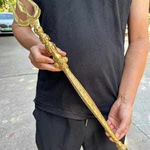 May include: A gold-coloured metal trident staff featuring a three-pronged top, a central column adorned with a skull and two sculpted faces, and a long, tapered handle decorated with intricate engraved patterns, ending in a small ornate finial.