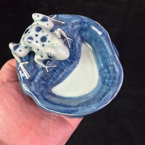 May include: A ceramic frog figurine with blue spots rests on a blue and white dish. The frog is white with blue spots and has detailed legs. The dish has a wavy design with a white interior and blue exterior.