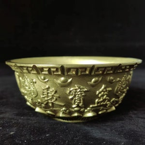 Könnte beinhalten: Eine goldfarbene Schale mit geprägten chinesischen Schriftzeichen und dekorativen Mustern. Die Schale hat einen breiten Rand und einen abgerundeten Boden. Die Zeichen und Muster sind erhaben und erzeugen eine strukturierte Oberfläche.