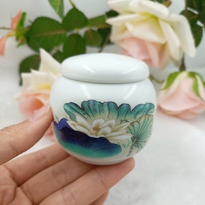 Puede incluir: Un pequeño tarro de cerámica blanco con tapa. El tarro está decorado con un diseño floral con una flor de loto blanca y hojas verdes y azules. El tarro está sostenido en una mano.