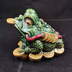 May include: A glossy green ceramic three-legged toad figurine featuring a textured, bumpy back, a red tongue, and a gold-colored coin held in its mouth. The toad is surrounded by and sits upon several gold-colored coins with embossed details.