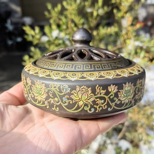 May include: A dark gray ceramic incense burner with a decorative lid. The burner features a floral design in yellow, green, and blue. The lid has a geometric pattern. The incense burner is round and held in a hand.