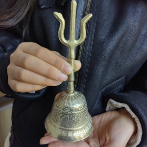May include: A brass bell with a trident-shaped handle. The bell has an aged, antique appearance with intricate etched designs around the base. The handle is a vertical rod with a trident at the top.