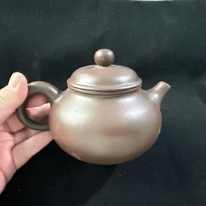 Op de afbeelding: Kleine, bruine keramische theepot met een afgeronde body, een gebogen handvat en een tuit. Het deksel heeft een kleine, ronde knop. De theepot heeft een gedempte bruine kleur en lijkt van klei te zijn.