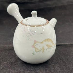 Puede incluir: Tetera de cerámica blanca con una ilustración de un gato durmiendo. La tetera tiene un cuerpo redondo, una tapa y un asa lateral. El gato es de color marrón claro y blanco, con orejas rosas y nariz rosa. La tetera tiene un esmalte craquelado.