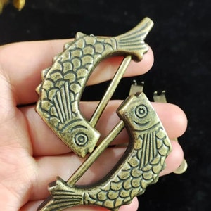 May include: Two antique bronze fish-shaped clasps. Each clasp features a fish design with detailed scales and fins. The clasps are connected by a metal rod, and the overall design has an aged, vintage appearance.