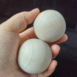 Puede incluir: Dos esferas de madera de color claro, sostenidas en una mano. Las esferas tienen una veta de madera natural y una forma lisa y redonda. La madera parece sin terminar, con un tono pálido y neutro. Las esferas miden aproximadamente 5 cm de diámetro.
