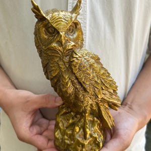 Könnte beinhalten: Eine goldfarbene Eulenfigur mit detaillierter Federstruktur, markanten Federohren und großen Augen. Die Eule sitzt auf einem strukturierten, felsartigen Sockel und weist eine metallische Oberfläche mit Licht- und Schatteneffekten auf.