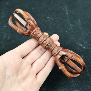 May include: A finely carved wooden vajra, a ritual object with a central shaft and ornate prongs. The brown wood displays detailed patterns and a symmetrical design. The vajra is held in a hand against a dark backdrop.