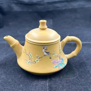 Może przedstawiać: Beżowy ceramiczny czajniczek z motywem ptaka i kwiatów. Czajniczek posiada uchwyt i dzióbek w stylu bambusowym. Ptak siedzi na gałęzi z delikatnymi kwiatami w odcieniach różu, zieleni i żółci.