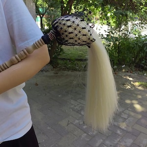 May include: A ceremonial item with a wooden handle and a decorative head covered in a black net. The head is attached to a long, flowing, light-colored tail. The handle has a decorative section with red, white, and blue stripes.