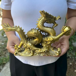 Peut inclure: Une sculpture de dragon en laiton doré avec des écailles complexes et une crinière fluide. Le dragon est posé sur des vagues stylisées, tenant une sphère dans sa griffe. La sculpture est de couleur dorée.