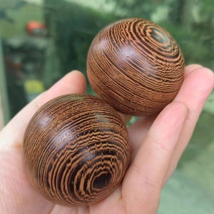 Può includere: Due sfere di legno con un motivo a strisce nei toni del marrone e del nero. Le sfere sono tenute in una mano, mostrando la venatura dettagliata del legno. Le sfere sembrano lisce e lucide.