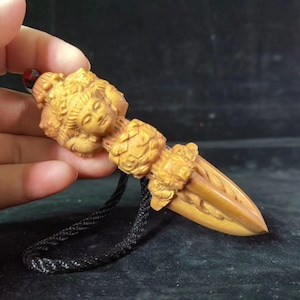 May include: A light brown wooden pendant carved into the shape of a multi-faced deity figure with intricate details, featuring a pointed base and a black braided cord attached to the top, accented by a small dark red bead at the very tip.