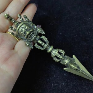 May include: A brass-coloured metal phurba ritual dagger featuring a detailed three-eyed deity face at the top, a central handle with intricate geometric carvings, and a sharp, pointed triangular blade at the bottom with a smooth finish.