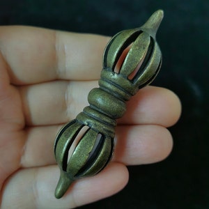 May include: A bronze-coloured metal vajra, a ritual object with a symmetrical design. The vajra features a central bulbous section and ends with stylized lotus-like shapes. The object is held in a hand against a dark background.