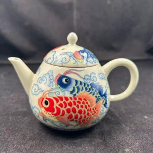 Könnte beinhalten: Cremefarbene Keramik-Teekanne mit Deckel und Henkel. Die Teekanne ist mit einem lebendigen Design aus roten und blauen Koi-Fischen verziert, die zwischen weißen Wolken schwimmen. Ausguss und Henkel sind farblich passend cremefarben.