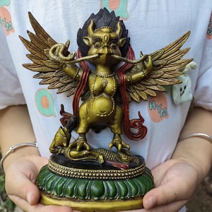May include: A gold-coloured statue of a winged deity with a bird-like head, horns, and a third eye, holding a snake in its beak. The figure stands on a green lotus base, trampling a smaller figure. It features red accents and detailed feathers.