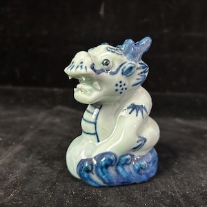 Puede incluir: Figura de dragón de cerámica en blanco y azul. El dragón tiene una cara detallada con la boca abierta y está decorado con detalles azules. La base presenta patrones de olas azules.