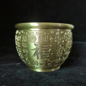 May include: A metallic gold-toned bowl with a rounded shape and a smooth rim. The exterior surface is decorated with repeating embossed Chinese characters and a geometric border pattern around the top edge, featuring a prominent central character.