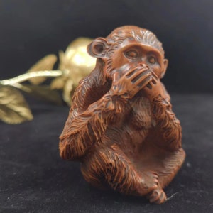 May include: A detailed wooden sculpture of a monkey covering its mouth with its hands. The monkey is carved with intricate details, showcasing fur and facial features. The sculpture is a warm brown color, with a dark background.