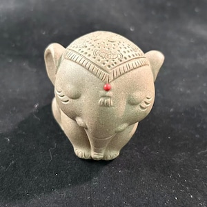 Puede incluir: Pequeña y detallada figura de un elefante sentado. El elefante es de un color verde-gris apagado con patrones grabados en la cabeza y un pequeño punto rojo en la frente. La trompa está enrollada y los ojos cerrados.
