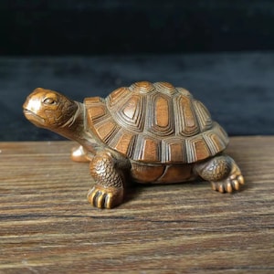 Op de afbeelding: Een gedetailleerd houten schildpadbeeldje met een getextureerd schild en gedetailleerde poten. De schildpad is gesneden uit lichtbruin hout en staat op een houten oppervlak. Het beeldje is een decoratief item.