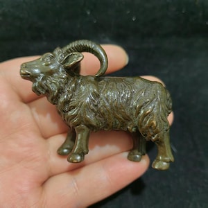 May include: A small, dark bronze-toned metal figurine of a ram, featuring detailed textured fur, a curved horn, and a bearded chin. The sculpture is cast in a standing position, showcasing a weathered, metallic finish with visible highlights.