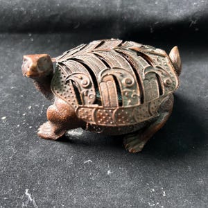 May include: A bronze-toned metal turtle figurine featuring a textured body and a removable, ornate shell lid with intricate cut-out patterns and engraved details. The turtle is depicted in a walking stance with a pointed head and a small tail.