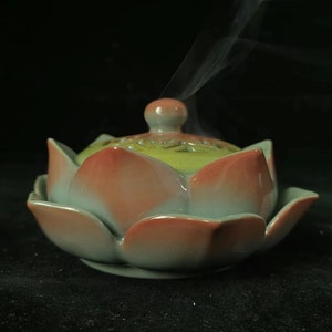May include: A ceramic incense burner in the shape of a lotus flower. The burner is primarily light green with peach-colored petals. Smoke rises from the top, indicating the incense is lit. The background is black.