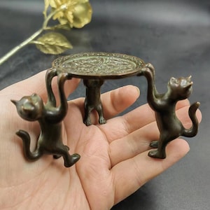 Könnte beinhalten: Ein bronzefarbener dekorativer Ständer mit Katzenmotiv. Zwei Katzenfiguren tragen eine runde, verzierte Tischplatte. Die Katzen befinden sich in einer spielerischen Pose und halten den Tisch über ihren Köpfen. Der Ständer dient wahrscheinlich zur Präsentation kleiner Gegenstände.