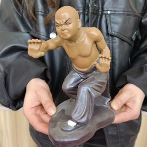 May include: A brown ceramic figurine of a bald man in a martial arts stance. The figurine is wearing dark gray pants and is posed on a dark gray base. The man's face has an angry expression.