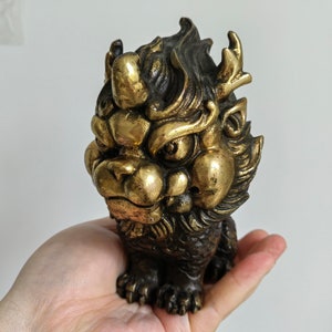 May include: A detailed bronze sculpture of a mythical creature with a lion-like face, horns, and intricate detailing. The statue has a gold and dark brown color scheme, with a textured surface. The creature is in a seated position.