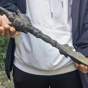May include: An antique bronze ceremonial scepter with detailed carvings. The scepter features dragon-like figures at the top and bottom, with intricate patterns along the shaft. The overall design suggests a historical or cultural significance. The scepter is held in both hands.
