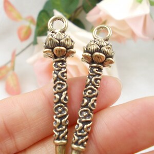 Puede incluir: Dos colgantes antiguos con forma de flor de loto de color latón. Cada colgante presenta una cabeza de flor de loto detallada con un lazo para colgar y un tallo decorativo con un patrón de remolino. Los colgantes son probablemente para la fabricación de joyas.