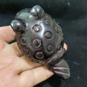 May include: A dark brown, carved frog figurine. The frog has a rounded body with circular indentations and prominent eyes. The sculpture is made of a dark, possibly polished material, and the details are well-defined.