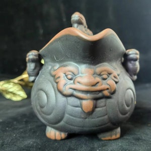 May include: A black ceramic pot with a smiling face design. The pot has a brown rim and accents, with two handles and three small feet. A small creature figure sits on the rim. The pot is likely used for decorative or functional purposes.