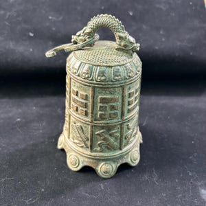 May include: Bronze-colored bell with a dragon-shaped handle. The bell features intricate carvings and a detailed design. The bell has a round base and a decorative pattern around the body. The bell is likely a decorative item or a collectible.