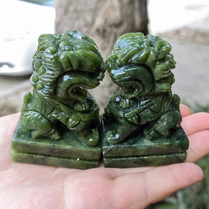 May include: Two jade green Foo Dog statues. Each statue is carved in detail, depicting a lion-like creature with a fierce expression. The statues are on a rectangular base. The jade has a glossy finish.